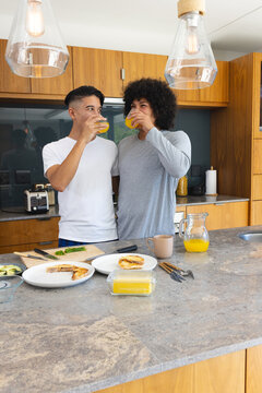 Drinking orange juice diverse male couple standing behind stone kitchen island, with toasted bagels