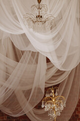 Two ornate chandeliers hang beneath flowing white fabric drapes against a brick wall. Concepts like luxury, celebration, event decor, or vintage, wedding invitations, interior design promotions.