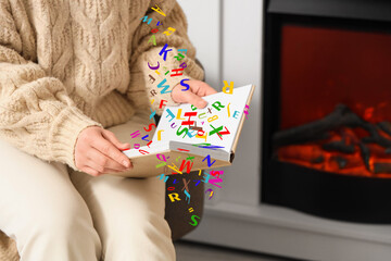 Young woman reading book in armchair near fireplace at home, closeup