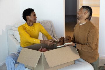 Diverse couple unpacking open box on light-colored sofa in living room, with brown leather shoes