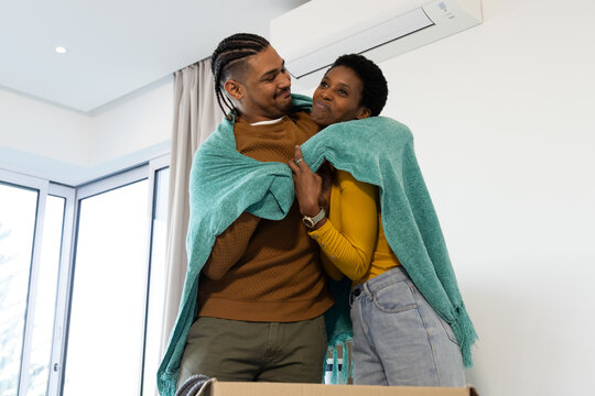 Sharing teal throw blanket, diverse couple standing at home near sliding windows, with moving box