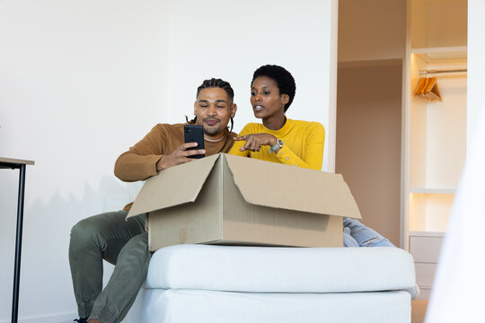 Diverse couple unpacking cardboard box on cushioned ottoman in sparse room, checking smartphone