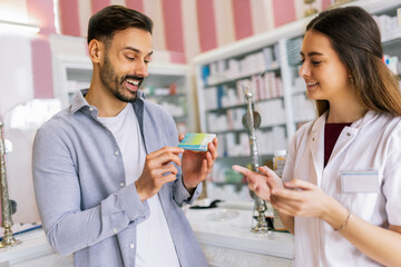 A pharmacy cashier is recommending medicine to a customer, while a professional pharmacist dispenses prescription medications in the pharmacy.