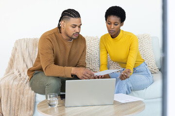 Reviewing paperwork Diverse couple sitting on sofa in living room, with laptop and documents