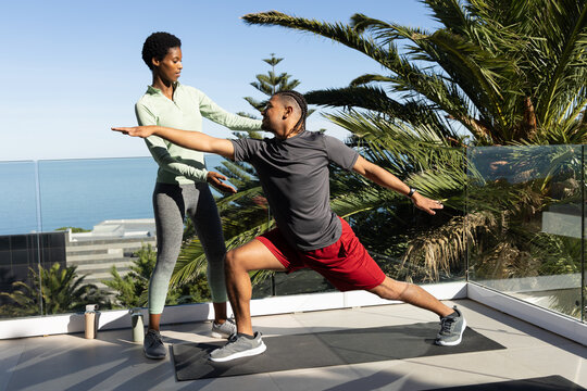 Diverse fitness duo practicing yoga lunge on sunlit ocean terrace, with black mat and water bottles