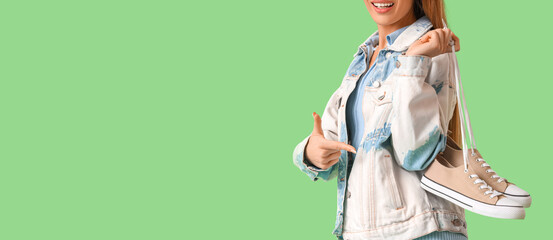 Stylish young woman pointing at sneakers on green background, closeup