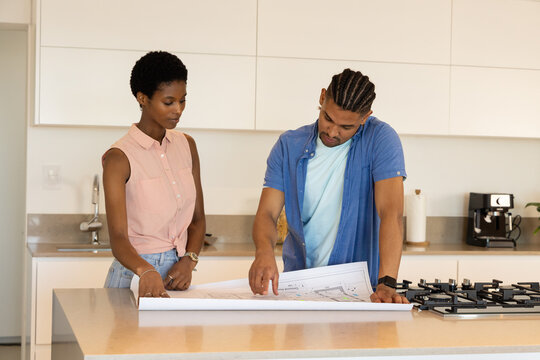 Examining blueprints, diverse couple planning layout on island countertop in modern kitchen
