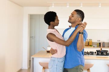 Dancing diverse couple holding hands by wooden kitchen island in white kitchen, with pasta jars