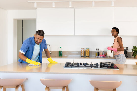 Diverse couple wearing gloves cleaning kitchen island in home kitchen, with spray bottle