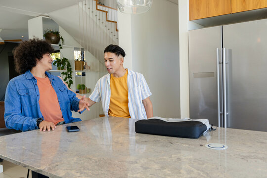 Laughing diverse male friends chatting at marble kitchen island, with smartphone and laptop bag