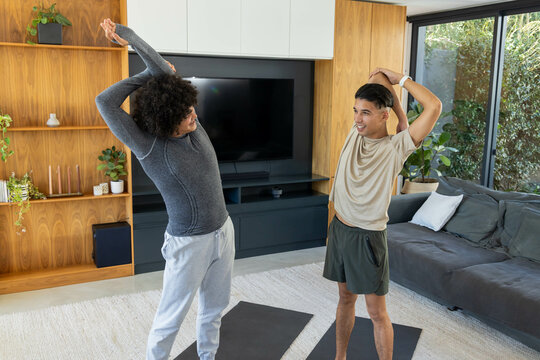 Diverse male friends stretching overhead in modern living room, with black yoga mats