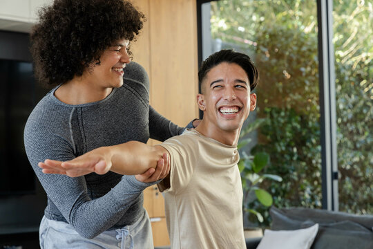 Assisting diverse male fitness trainer and client stretching arms in modern living room, with sofa