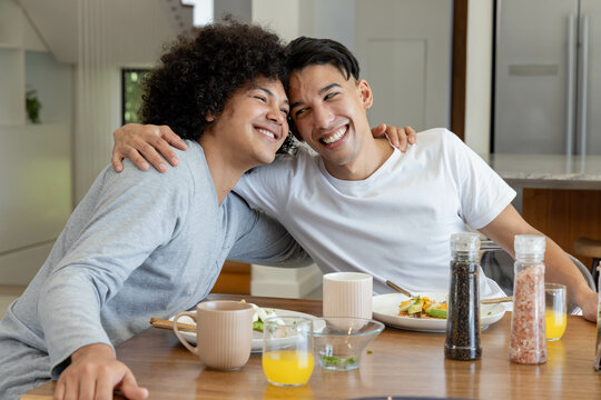 Smiling diverse male friends hugging at modern kitchen dining table, with glass of orange juice - Powered by Adobe