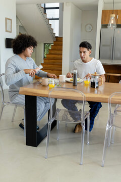Sitting Diverse father and son eating breakfast at open-plan kitchen, with French press and salt
