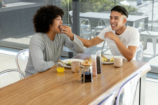 Laughing Diverse male friends sharing breakfast in dining nook, with wooden table and coffee maker - Powered by Adobe