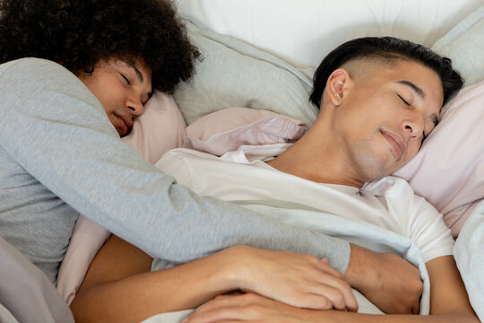 Cuddling diverse male partners lying on bed at home, with pastel pillows, blankets and sheets - Powered by Adobe