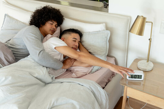 Cuddling Diverse male couple lying on bed in modern bedroom, with digital alarm clock