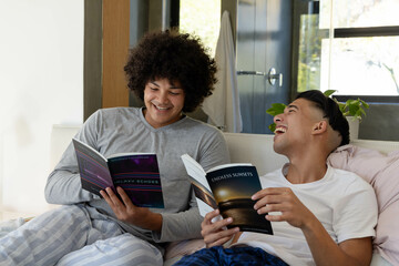 Diverse male friends laughing and reading glossy magazines on sofa at home, with potted plant