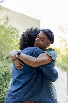 Hugging diverse father and son sharing embrace on outdoor walkway beside building, with smartwatch