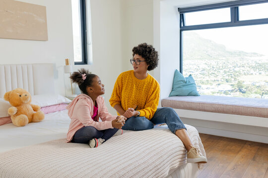 Mother and daughter holding hands sitting on bed in bedroom, with teddy bear and hillside view