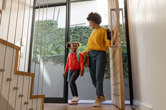 Smiling Diverse mother and daughter standing in front doorway, with smartphone and teal backpack