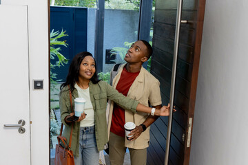 African American couple entering modern entryway, carrying coffee cups, tote bag and smartphone