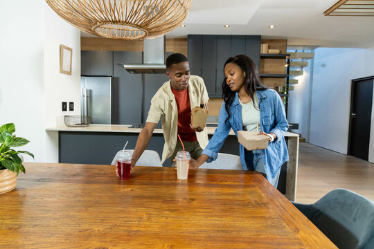 African American couple placing takeout boxes and cups on table in modern kitchen, pendant light - Powered by Adobe