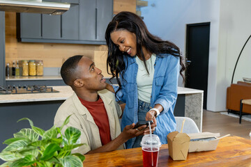 Holding smartphone African American couple leaning at kitchen table, with red drink and takeout