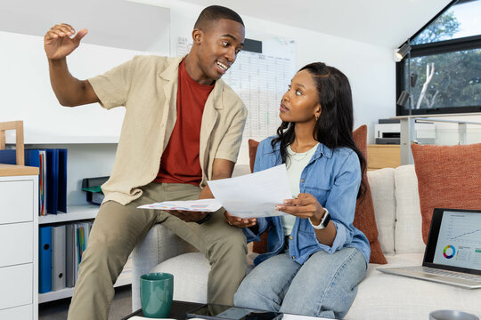 African American couple analyzing printed documents in home office, with coffee mug and laptop