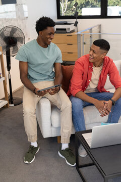 Chatting diverse male friends lounging on light gray couch in living room, with laptop and tablet