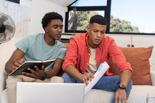 Studying diverse male friends reviewing papers on living room couch, with laptop and whiteboard