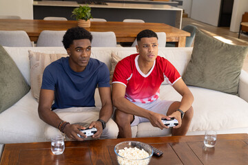 Playing Diverse male friends gripping white game controllers on sofa in living room, with popcorn