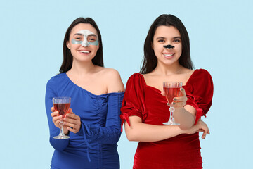 Beautiful young women in stylish dresses with facial masks drinking wine on color background