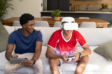 Wearing VR headset diverse male friends sitting on couch in living room, with tablet and controller