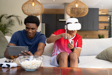 Diverse male friends using VR headset and browsing tablet on sofa in living room, with popcorn