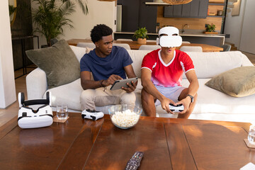 Gaming diverse male friends wearing VR headset and holding tablet on couch in modern living room