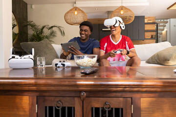 Laughing Diverse male friends using tablet and VR headset in modern living room, with popcorn