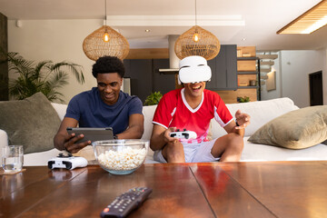 Playing VR games diverse male friends sitting on sofa in living room, with controllers and headset