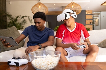 Sitting Diverse male friends using tablet and wearing VR headset while gaming on sofa, with popcorn