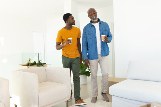 Walking African American father and son carrying coffee cups in bright living room, with moving box