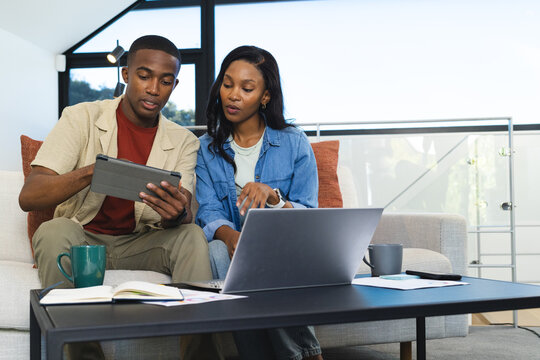 Working African American couple using tablet on living room sofa, with laptop and notebooks