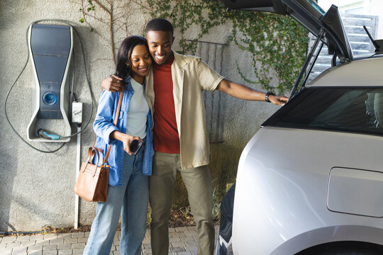 African American couple hugging on driveway, opening EV hatch at charging station, copy space