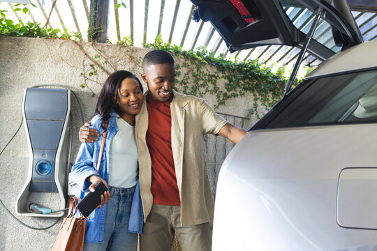Standing African American couple examining white electric hatchback under pergola, with charger