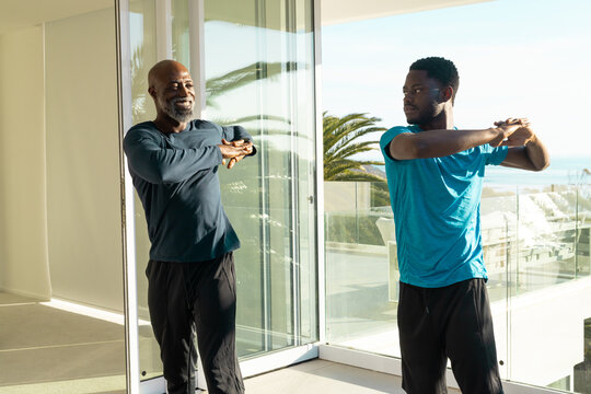Stretching African American father and son pulling arms across chest in home, with glass doors