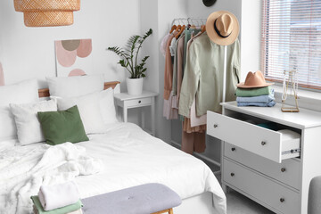 Comfortable bed, chest of drawers and rack with stylish clothes in interior of bedroom