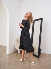 Blonde woman in a black polka dot dress stands confidently near a large mirror, with a neutral...