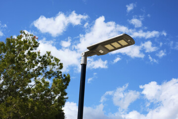 Modern solar-powered streetlight against vibrant blue sky with white clouds and green foliage