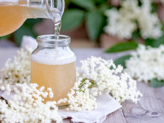 A liquid is being poured into a glass jar on a light wooden surface, with a bunch of white flowers...