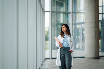 Female doctor walking in modern hospital corridor holding patient chart