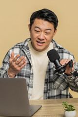 Happy excited blogger Asian man at table recording podcast giving workshop, webinar. Chinese guy influencer speaking into microphone and laptop webcam, making video call discussing on beige background